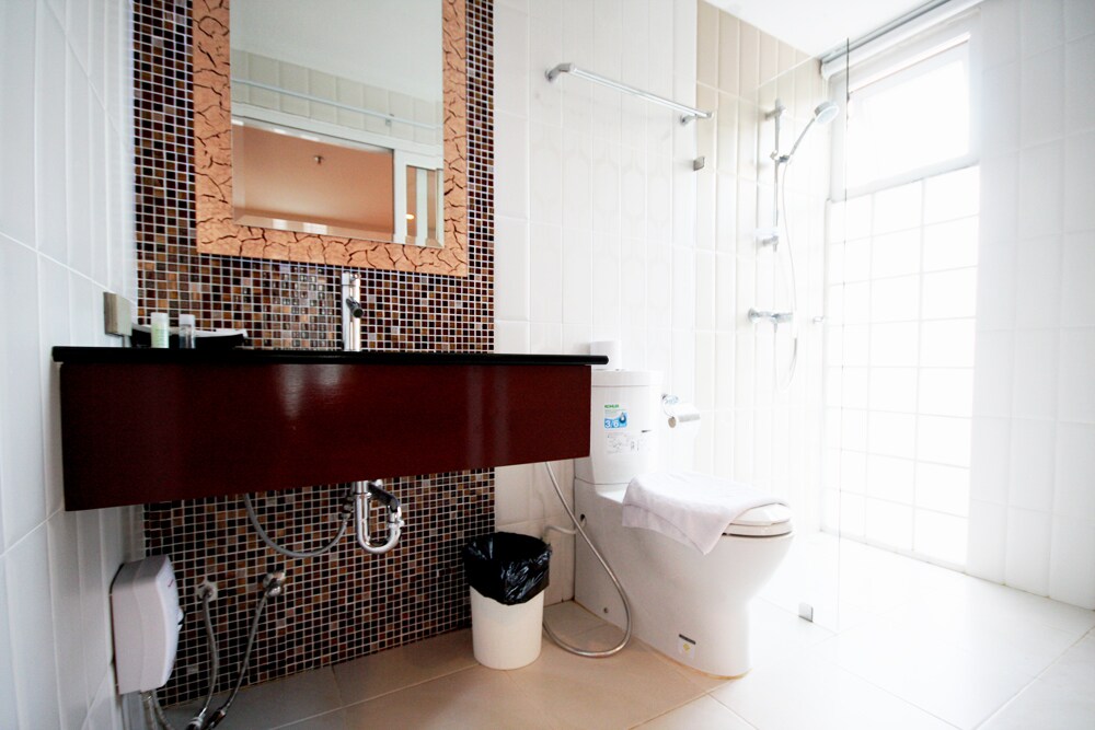 A modern bathroom features a stylish mosaic-tiled wall and a large mirror above a dark wooden vanity. A white toilet is positioned next to a glass shower space with rainfall showerhead, allowing for a clean and well-lit environment.