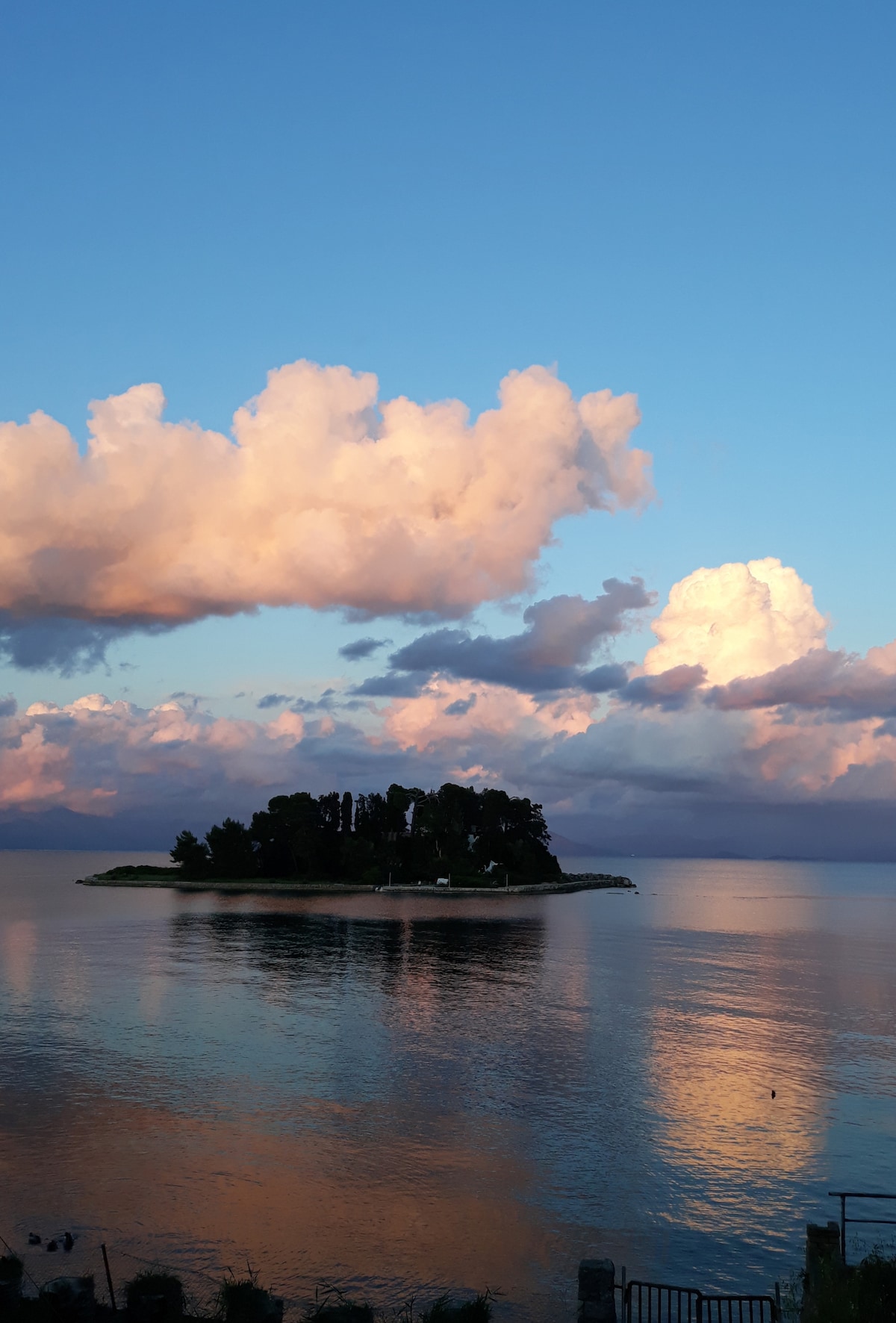 A scenic view of a small island surrounded by calm waters, reflecting the colors of the sky. Fluffy clouds in shades of pink and white are scattered across a blue background, creating a serene atmosphere. The tranquility of the water contrasts with the landmass, enhancing the natural beauty of the location.