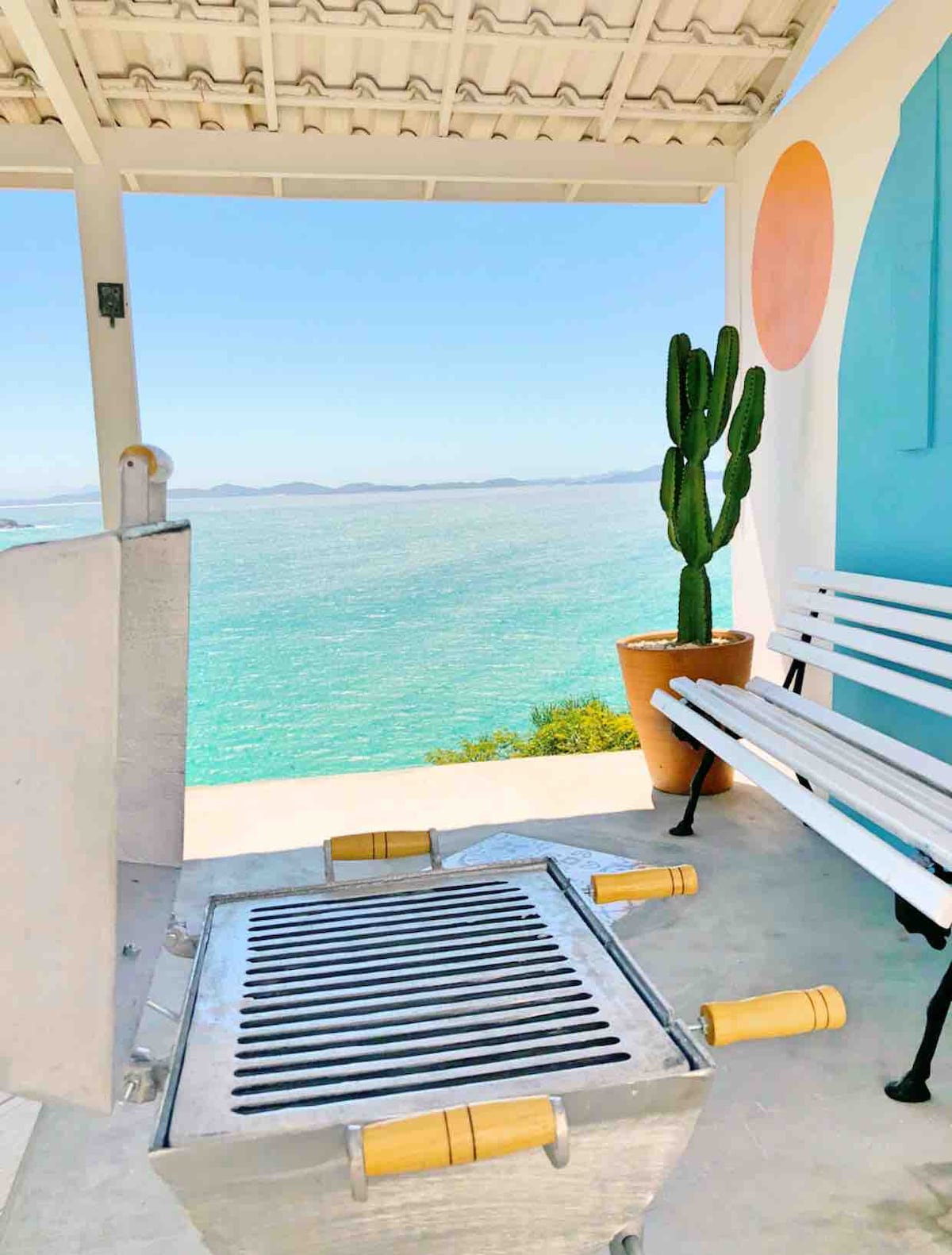 An outdoor area features a portable grill positioned on a concrete platform, overlooking the turquoise sea. A white bench sits against the wall, complemented by a large potted cactus. The space is bright, with clear views of the water and distant islands.