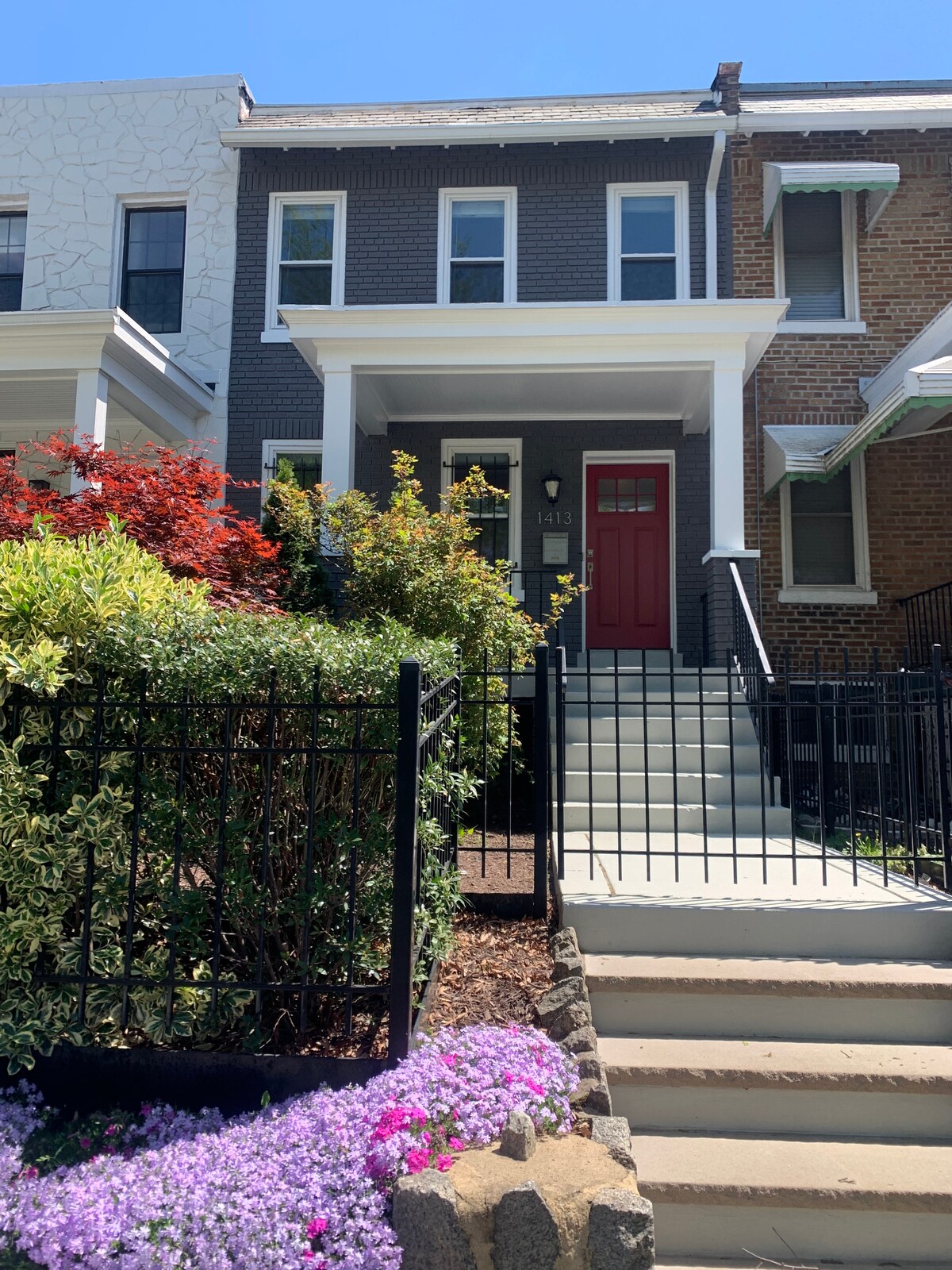 A charming exterior view of a well-maintained home, featuring a bright red front door and tasteful landscaping. Colorful flowers are in bloom at the base of the stairs, which lead to the entrance. A black fence outlines the front garden, providing a welcoming touch.