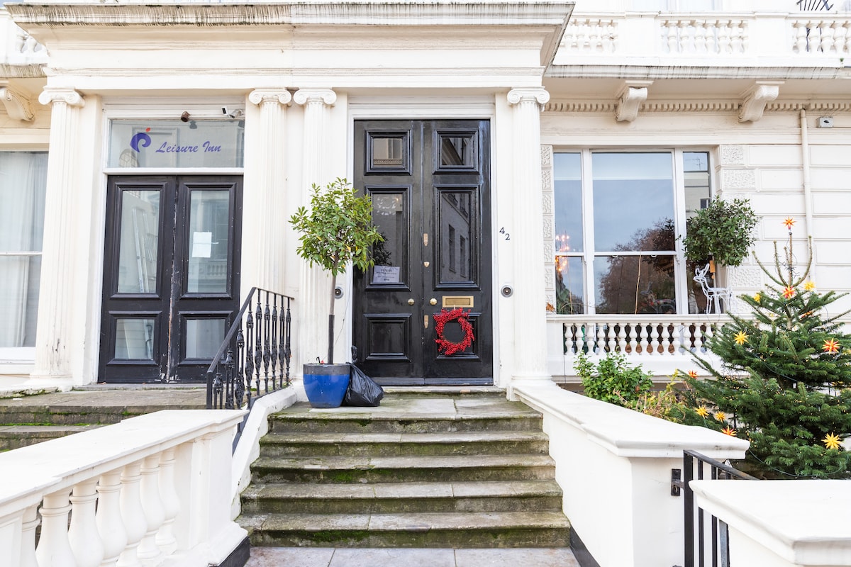 The entrance to a historic grade two listed building is showcased, featuring a set of stone steps leading to a dark wooden door adorned with a red wreath. Flanking the doorway are potted plants, and a small, decorated tree is visible nearby.