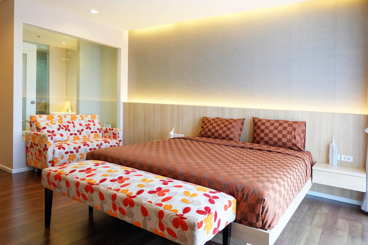 A modern bedroom features a spacious bed with a patterned brown coverlet, flanked by two decorative cushions. A colorful seating area with floral-patterned chairs is present, accompanied by a bench adorned with matching fabric. Warm lighting highlights the textured wall behind the bed.