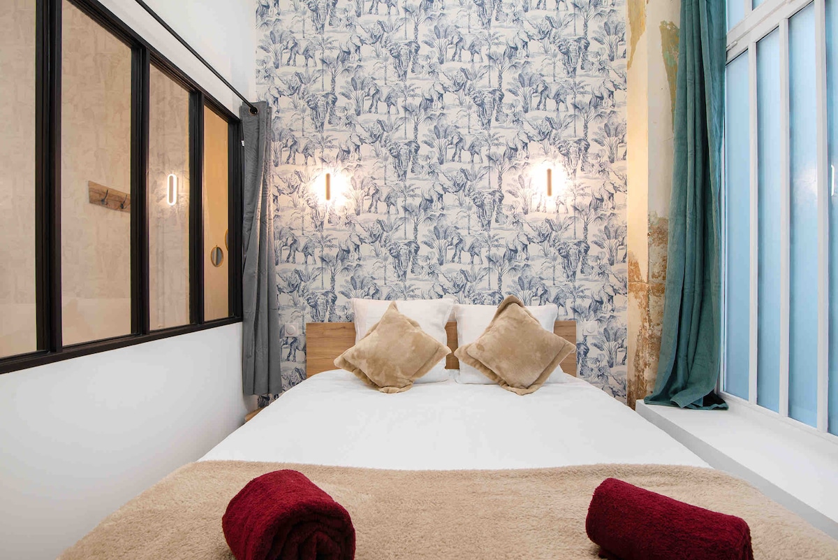 A bright bedroom features a spacious bed adorned with white linens and plush pillows. Soft textures are complemented by decorative cushions. A floral wallpaper adds visual interest, while natural light filters through large windows, illuminating the neutral tones of the space.