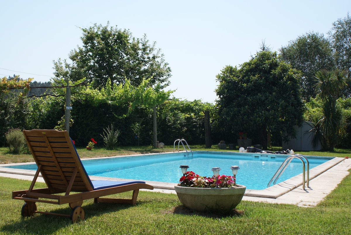 A swimming pool is surrounded by greenery, with clear water reflecting the sky. A lounge chair is positioned nearby on a grassy area, while flower pots with vibrant plants add color to the space. Lush trees provide shade and privacy around the pool.