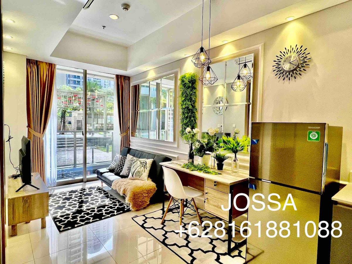 A stylish living area features a black and white patterned rug and a modern sofa, complemented by natural greenery on the wall. Soft light enters through large glass doors that lead to a swimming pool view. A dining table and a sleek refrigerator enhance the comfort.