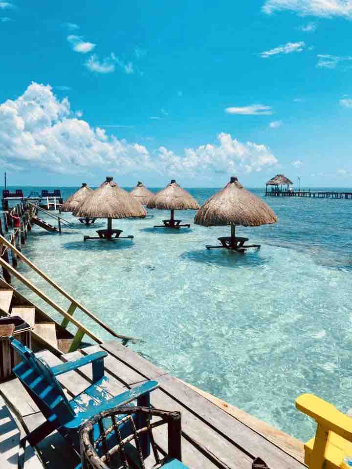 Shady thatched-roof palapas are positioned over calm, clear waters, offering relaxation spots along a wooden deck. Brightly colored chairs are visible on the deck, inviting guests to enjoy the vibrant seaside environment under a clear blue sky.