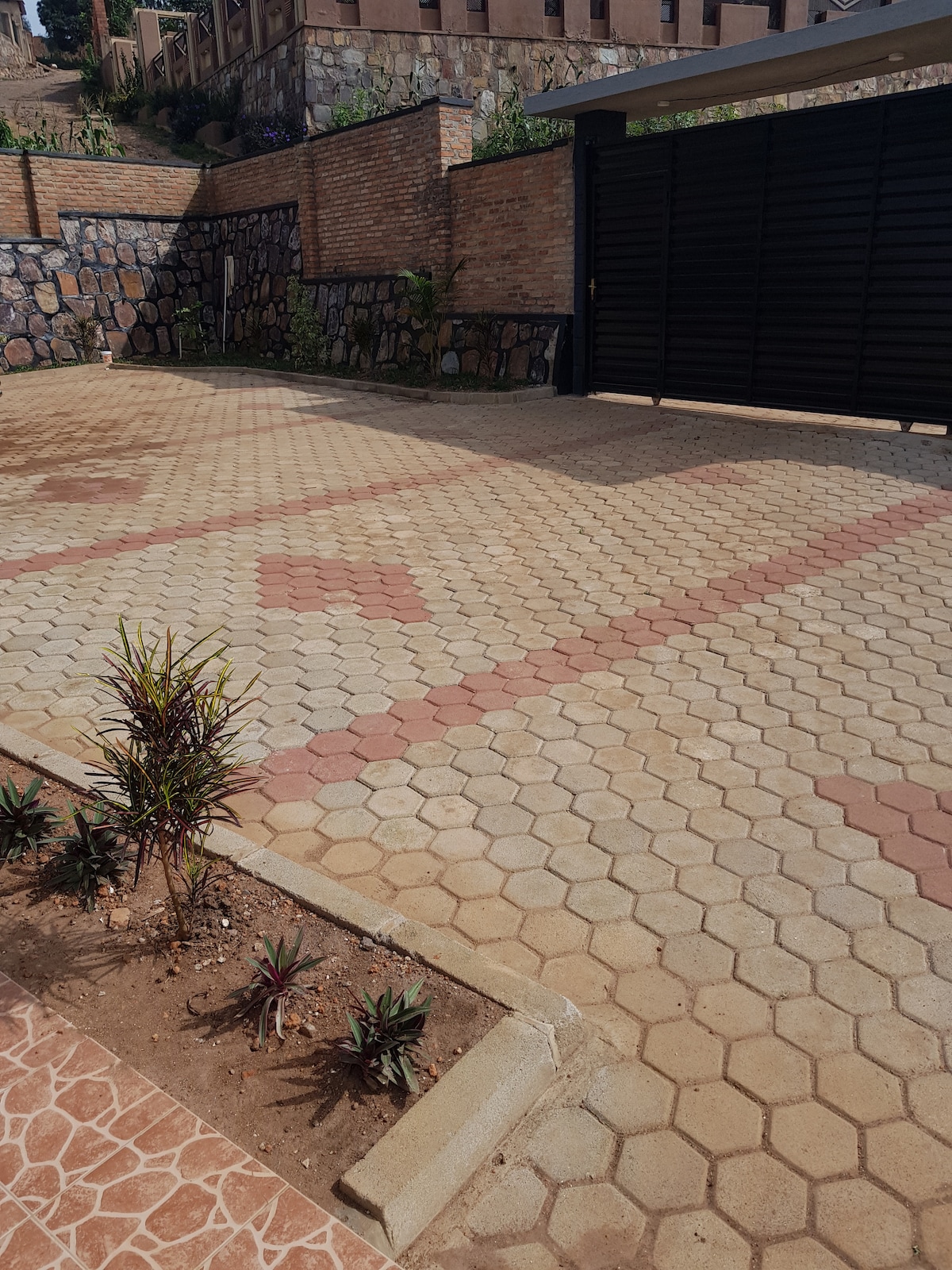 A spacious driveway is paved with hexagonal stones in alternating colors, creating a geometric pattern. Decorative plants are positioned along the edges, contributing to the overall appearance. The area is enclosed by a stone wall and a closed gate is visible in the background.