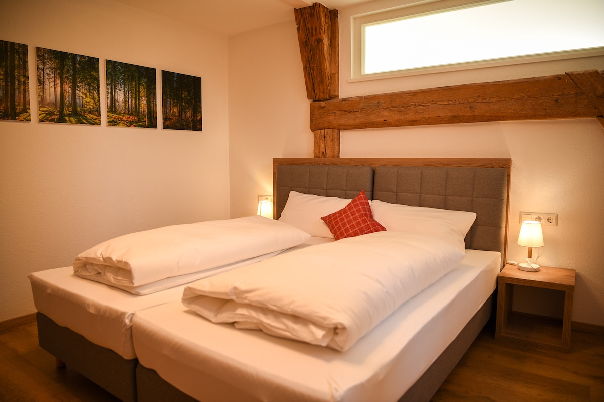 A bright bedroom features a spacious bed with two separate mattresses topped with white bedding. Wooden beams and a large window enhance the natural light. Two bedside tables with lamps provide illumination, while three landscape paintings of a forest are displayed on the wall.