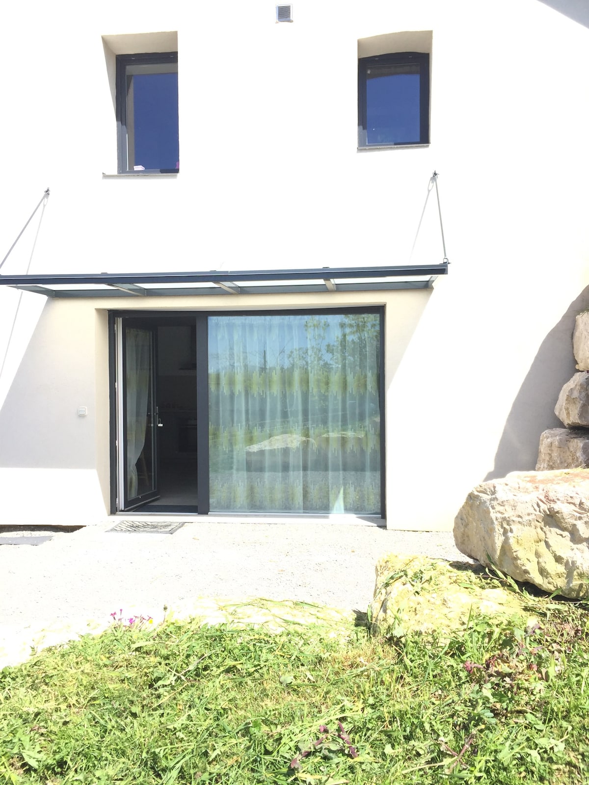The entrance to the flat is framed by a modern overhang, featuring a large glass door that opens onto a well-lit interior. Two small windows above allow natural light to filter in. A gravel pathway leads to the door, complemented by a patch of green grass and wildflowers.