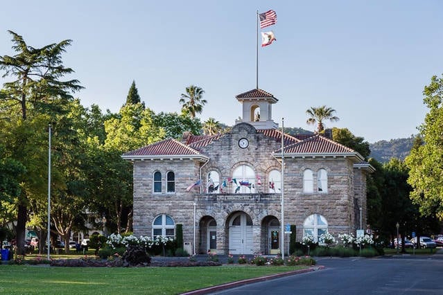 A historic stone building features a red-tiled roof and a central clock tower. Lush greenery surrounds the structure, with palm trees and vibrant flowering plants enhancing the scenery. The façade showcases arched windows and multiple flags displayed prominently.