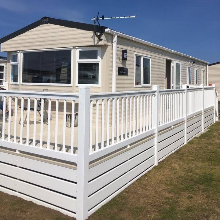 A modern caravan with a light exterior stands under clear skies. A spacious deck encircled by white railings features outdoor seating, while multiple large windows enhance natural light. The manicured grass area around the caravan contributes to the inviting outdoor setting.