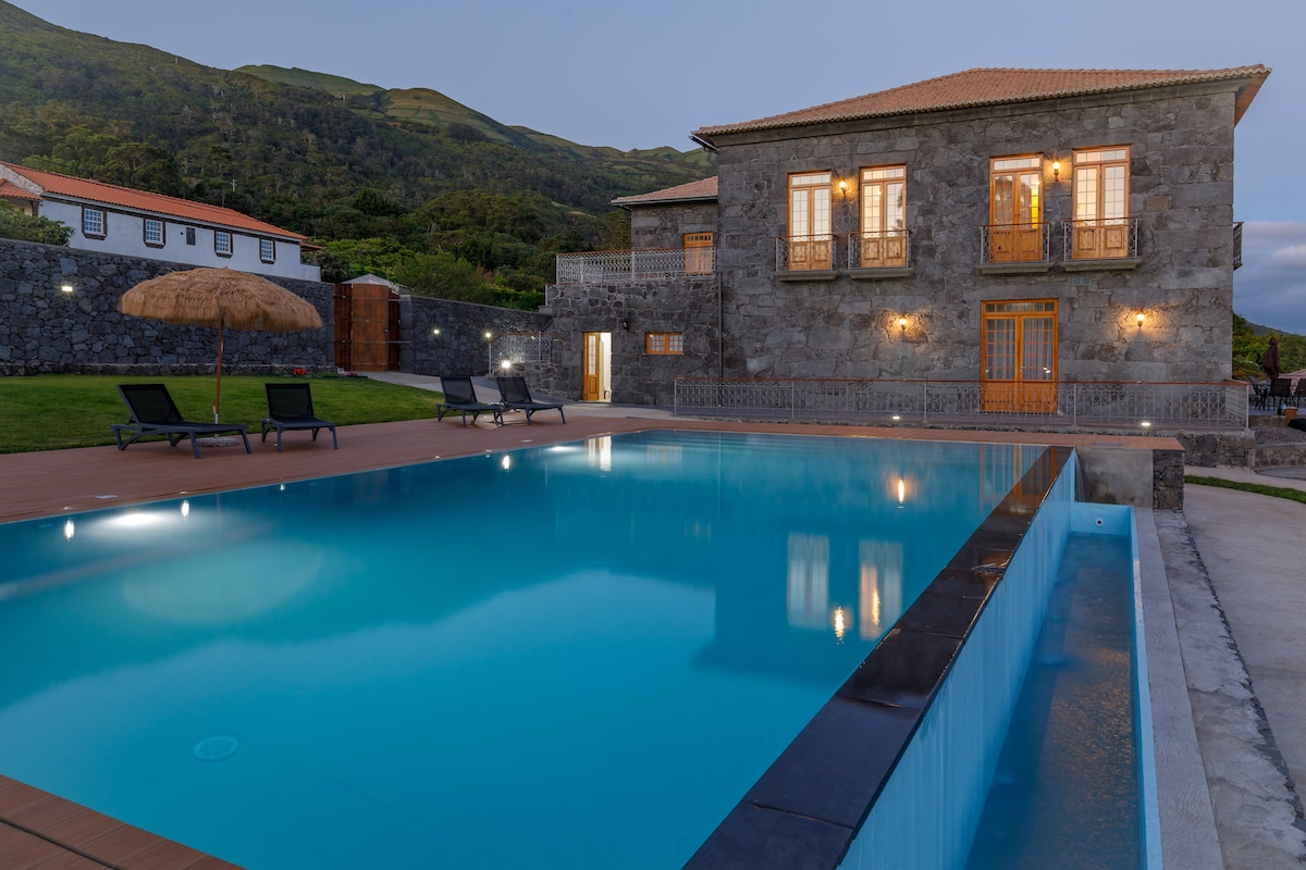 A stone house is seen beside a large swimming pool, with two lounge chairs positioned nearby. The property is set against a backdrop of lush mountain scenery, and warm lighting is visible through the windows, creating a welcoming ambiance in the evening.
