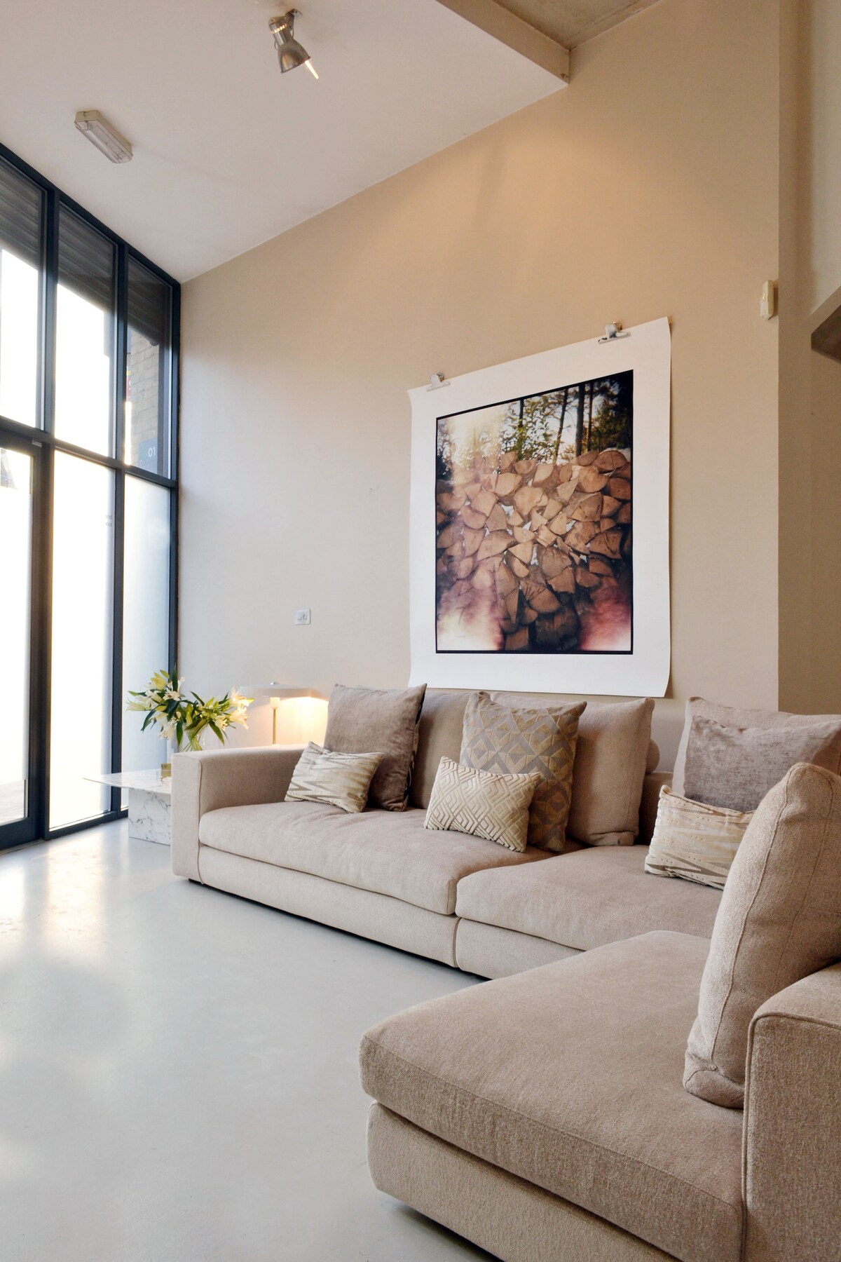 A spacious living area is highlighted by large windows allowing natural light to fill the room. A light-colored sofa is adorned with various cushions, and a piece of art featuring natural stone is displayed prominently on the wall. A small table rests beside the sofa.