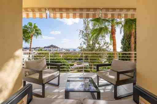 A comfortable outdoor seating area is presented, featuring two armchairs and a coffee table. Sunlight filters through the protective awning, highlighting the distant sea view and greenery surrounding the space.