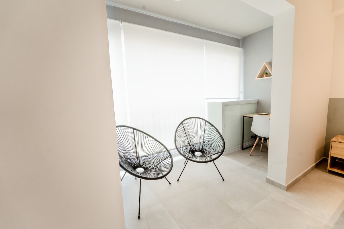 Two contemporary Acapulco chairs are positioned near large, light-filtering windows that create a calming atmosphere. A small desk and a minimalist chair are visible, complementing the clean lines and neutral color palette of the space. The flooring is tiled, enhancing a modern aesthetic.