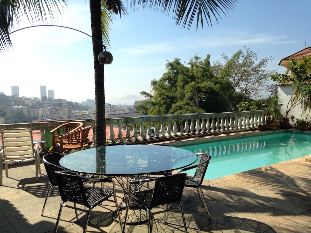 A spacious terrace is presented, featuring a circular glass table surrounded by black wicker chairs. A refreshing swimming pool is visible in the foreground, with lush greenery and a scenic view of the city skyline in the background, complemented by a clear blue sky.
