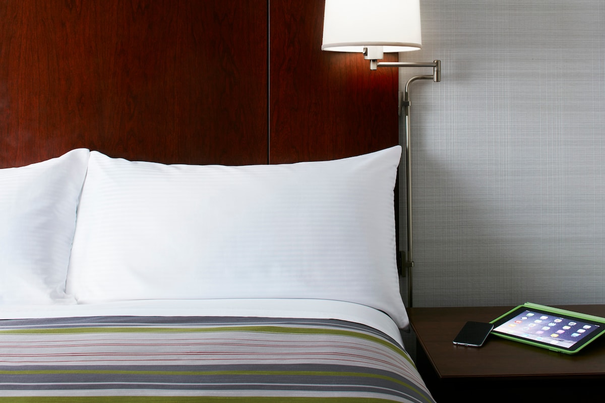 A cozy sleeping area features a spacious bed with crisp white bedding and a striped blanket. A sleek lamp beside the bed provides soft illumination. An equipped nightstand holds an electronic tablet and a smartphone, offering convenience for work or leisure.