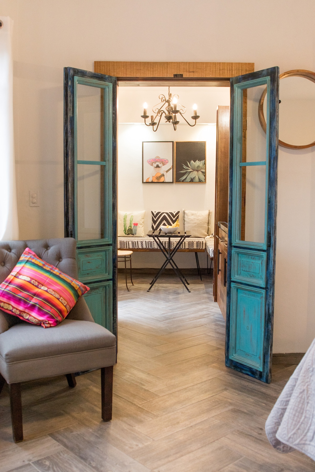 An inviting entryway is framed by vintage blue doors, leading to a cozy seating area. A small round table with two chairs sits beneath a stylish chandelier, while colorful wall art and decorative throw pillows add a touch of warmth to the space.