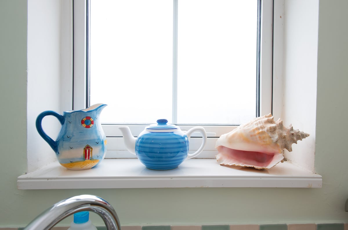 A windowsill displays three decorative items: a colorful pitcher with a beach scene, a blue teapot, and a large conch shell. Natural light filters through the window, enhancing the serene ambiance.