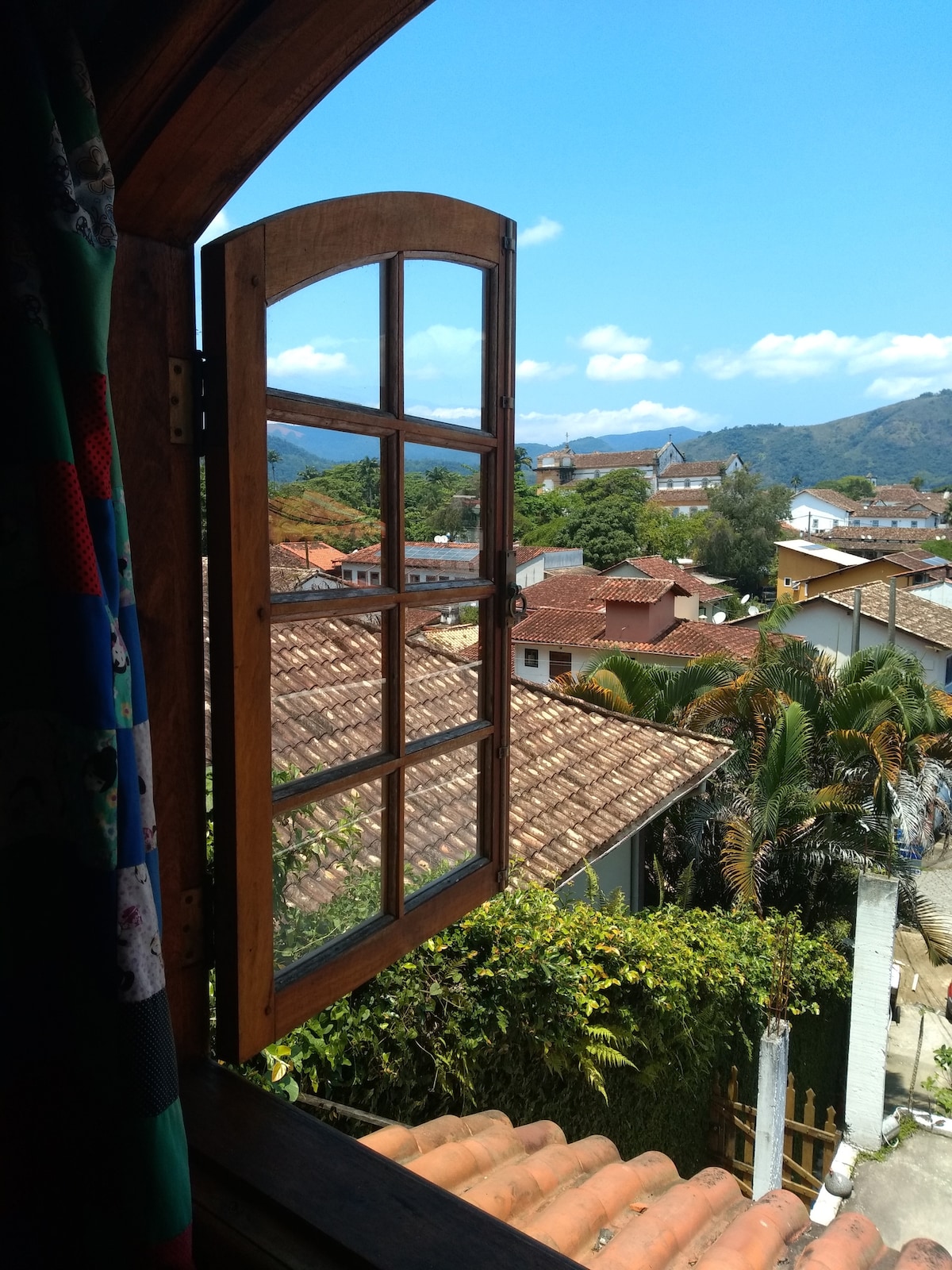 A wooden window with multiple panes opens to reveal a scenic view of the surrounding area. The landscape includes terracotta rooftops, lush greenery, and distant mountains under a clear blue sky. Sunlight brightens the scene, enhancing the vibrant colors of the neighborhood.