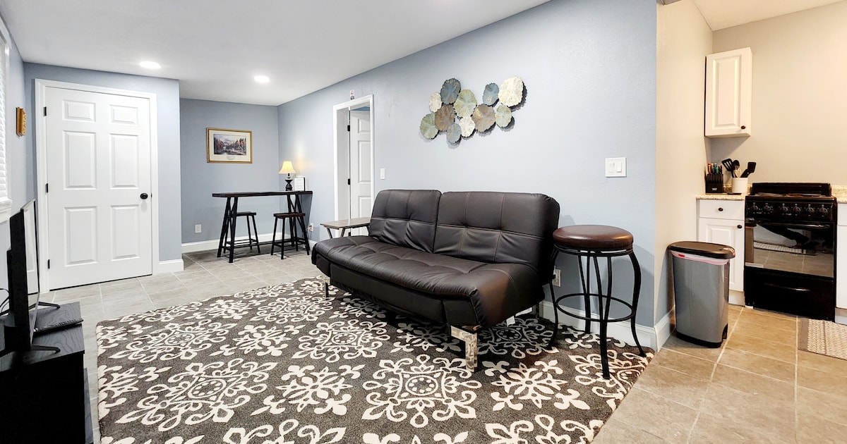 A living area features a dark sectional sofa positioned on a patterned rug. A small dining table with chairs is visible, along with a kitchen area containing a stove and countertop. Light-colored walls and recessed lighting create a bright and inviting space.