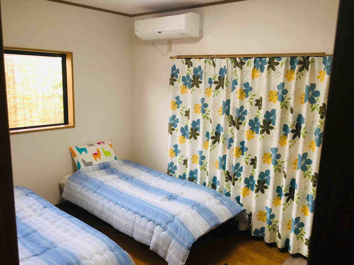 A room is equipped with two beds, each adorned with light blue bedding. A decorative cushion with a colorful design sits on one bed. A window with a wooden frame allows natural light to enter, while a floral curtain with blue and yellow flowers adds a touch of color.