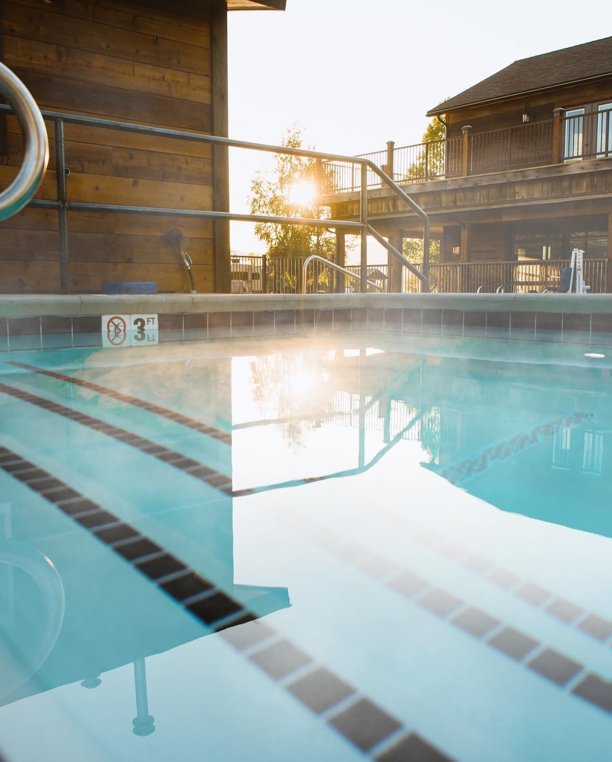 A heated outdoor pool is captured in the early morning light, with steam rising from the surface. The surrounding wooden structure is visible, and a gentle glow from the rising sun reflects in the water, enhancing the tranquil ambiance.