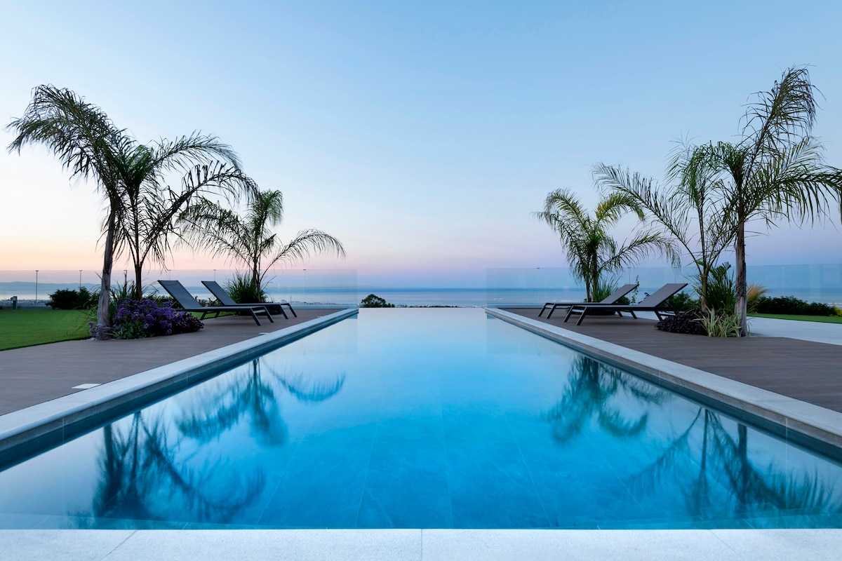 An infinity pool stretches invitingly towards the horizon, reflecting the soft hues of a rising sun. Palm trees frame the poolside, standing beside comfortable loungers. The calm waters and coastal landscape create a serene atmosphere, perfect for relaxation and enjoying panoramic ocean views.