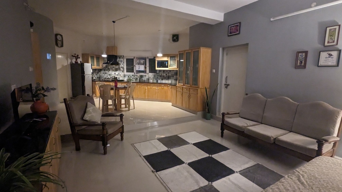 An open living area features a comfortable seating arrangement with light-colored sofas and a patterned area rug. The modern kitchen is visible in the background, showcasing wooden cabinetry and a dining table arranged with chairs. Natural light fills the space, creating a bright ambiance.