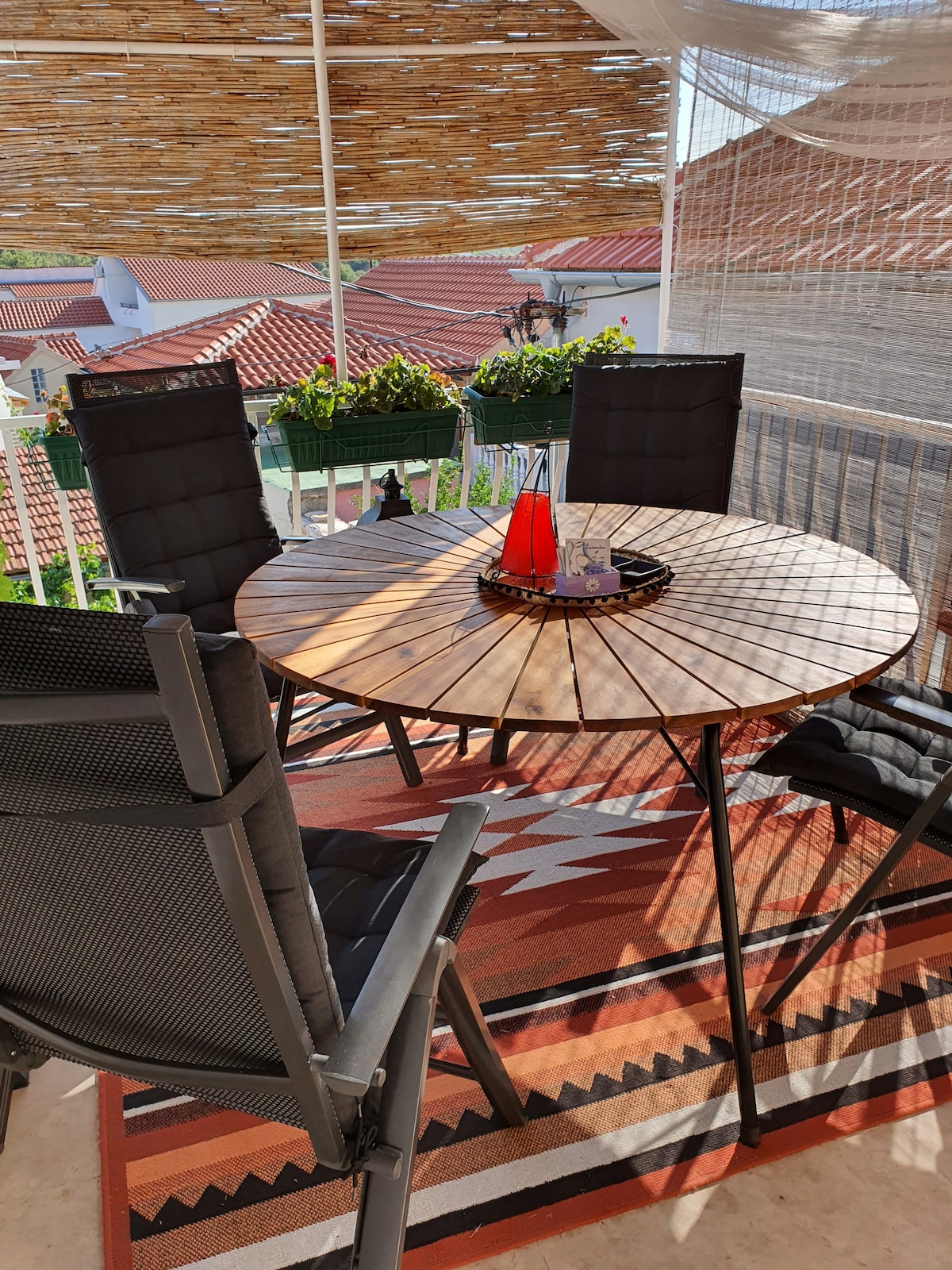 A circular wooden table is set at the center of a terrace, surrounded by four black chairs. The area features a patterned rug, with sunlight creating a play of shadows. Potted plants add greenery, enhancing the outdoor dining experience.