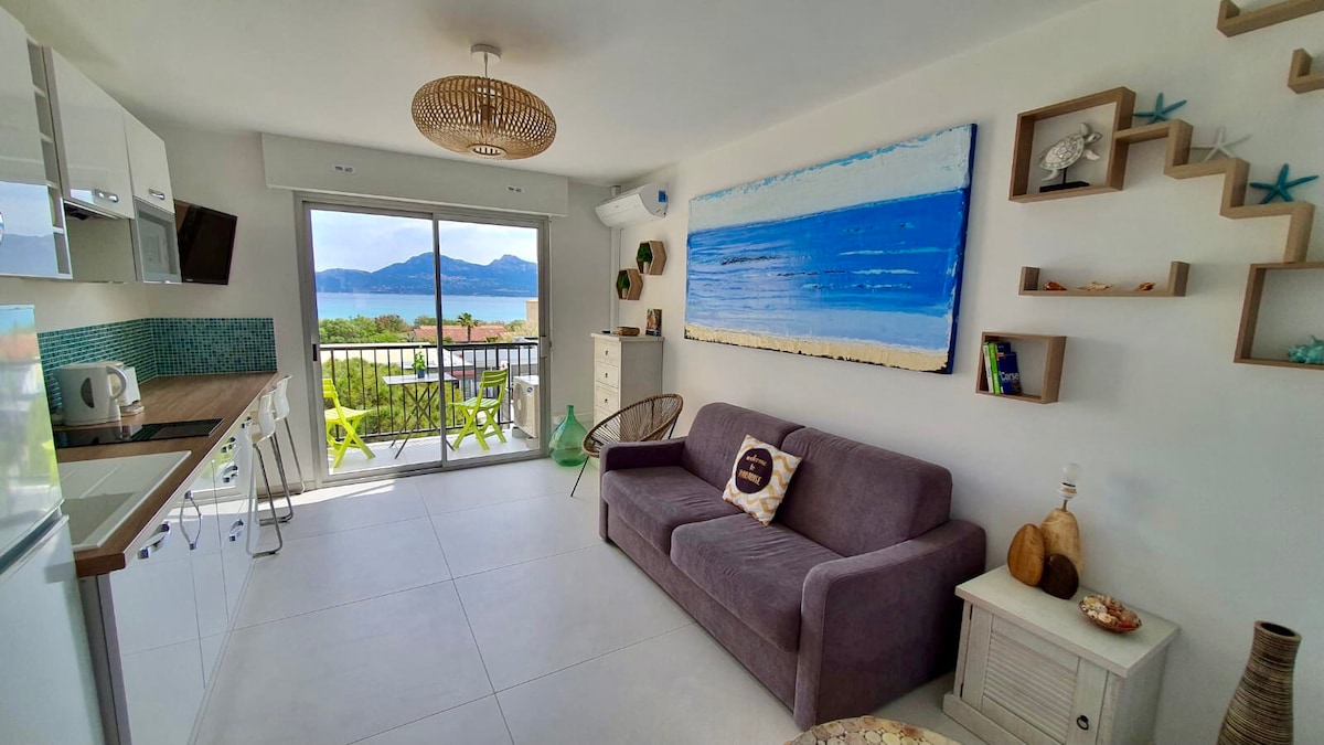 A modern living space features a spacious sofa facing large glass doors that open to a balcony with ocean views. Light wood shelving and a vibrant wall art piece enhance the room's aesthetic. The kitchen area is equipped with sleek white cabinets and appliances.