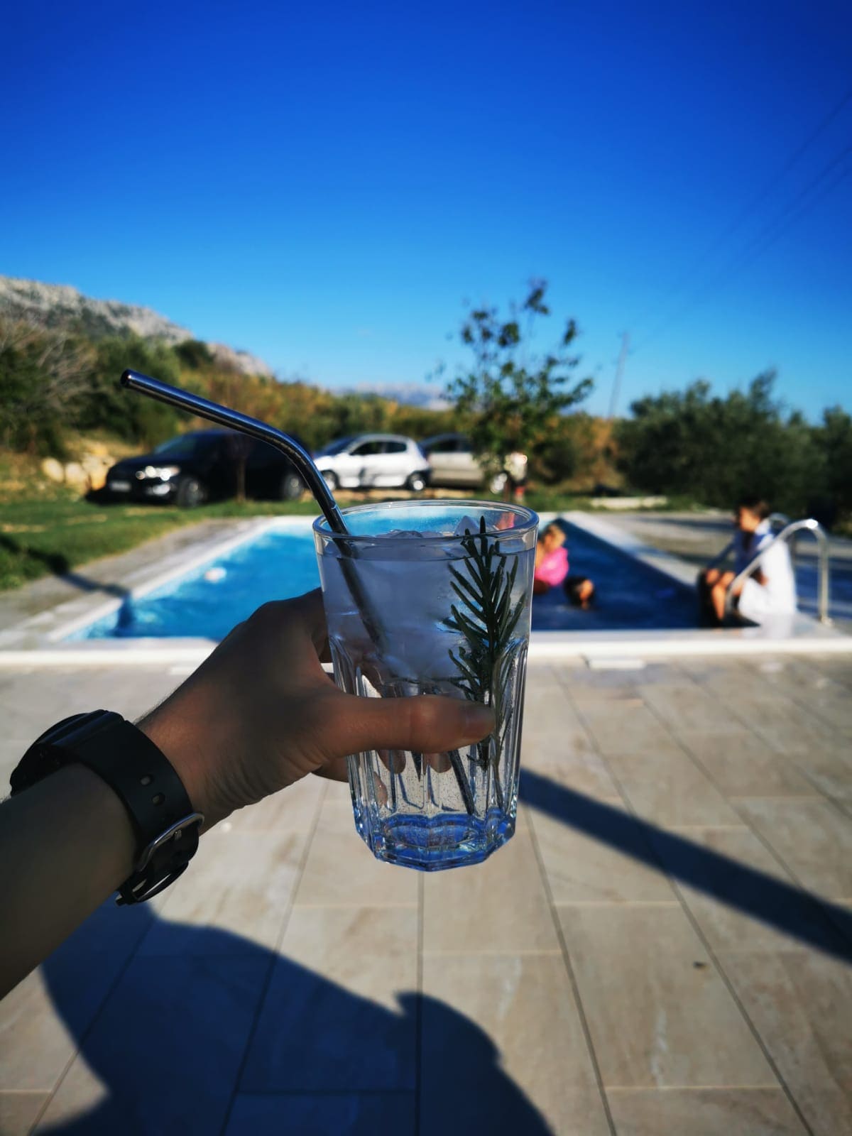 A clear glass with a sprig of rosemary is held in the foreground, showcasing a refreshing drink. In the background, a shared pool can be seen with people enjoying leisure time under the bright blue sky, surrounded by lush greenery.