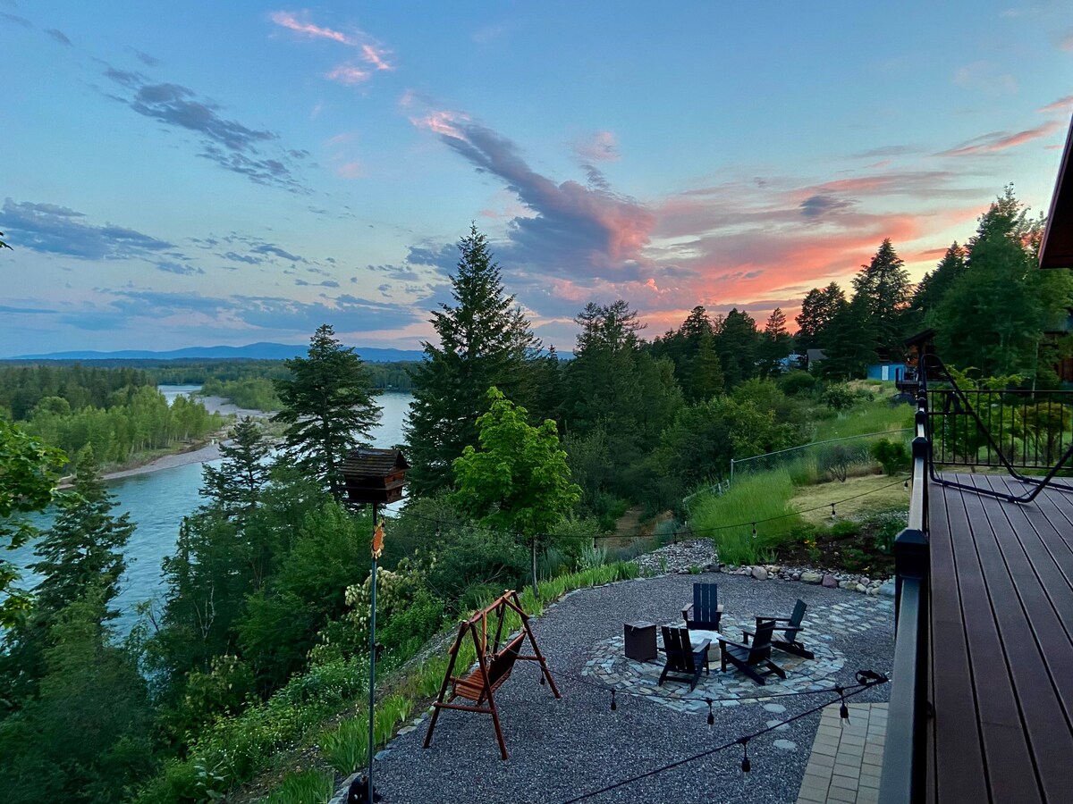 The outdoor patio area is equipped with a stone fire pit surrounded by several chairs, providing a perfect spot to enjoy the serene views of the Flathead River and surrounding greenery. The sky reflects soft hues during sunset, while trees frame the landscape.