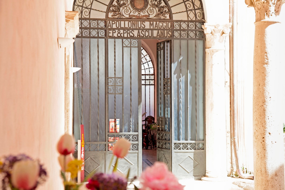 An elegant entrance features a large, wrought iron gate with decorative patterns at the forefront. Natural light streams through the open gate, illuminating the corridor beyond, which is characterized by stone columns and historical architecture, inviting guests to explore further.