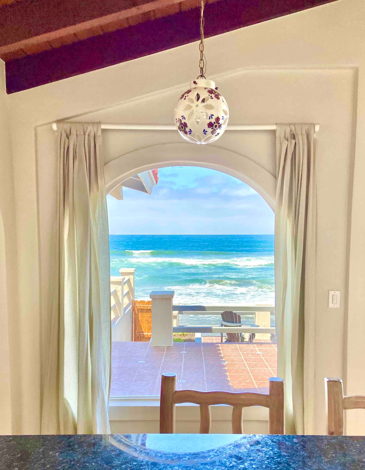 An arched window frames a stunning view of the Pacific Ocean, with sunlight reflecting off the waves. Light-colored curtains gently drape beside the window, while a wooden table and chairs are positioned in the foreground, inviting relaxed indoor gatherings.