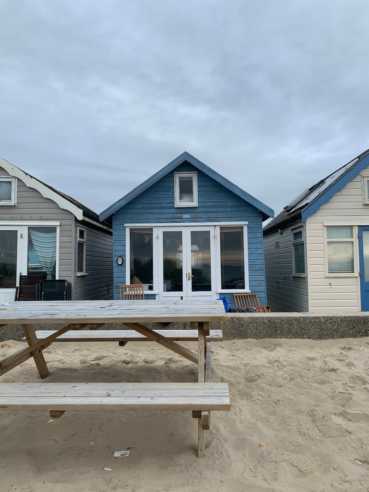 The beach hut, painted in a calming blue, stands centrally between two other huts with distinctive colors. A picnic table is positioned on the sandy foreground, providing a space for outdoor gatherings. The sky overhead is overcast, creating a serene coastal atmosphere.