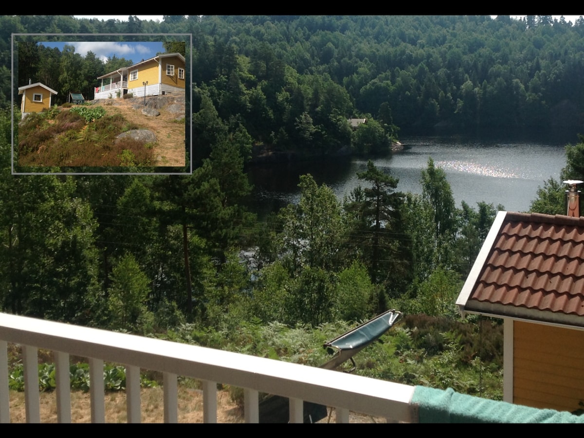 A serene view is presented from the terrace showing a calm lake surrounded by lush greenery. The main cottage with an annex is visible on the hillside, blending naturally with the landscape. Gentle slopes lead down to the water, enhancing the peaceful atmosphere.