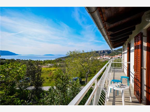 A balcony is presented with two blue chairs and a small table, offering views of the sea and mountains. Sunlight brightens the scene, highlighting the lush greenery and coastal landscape in the distance.