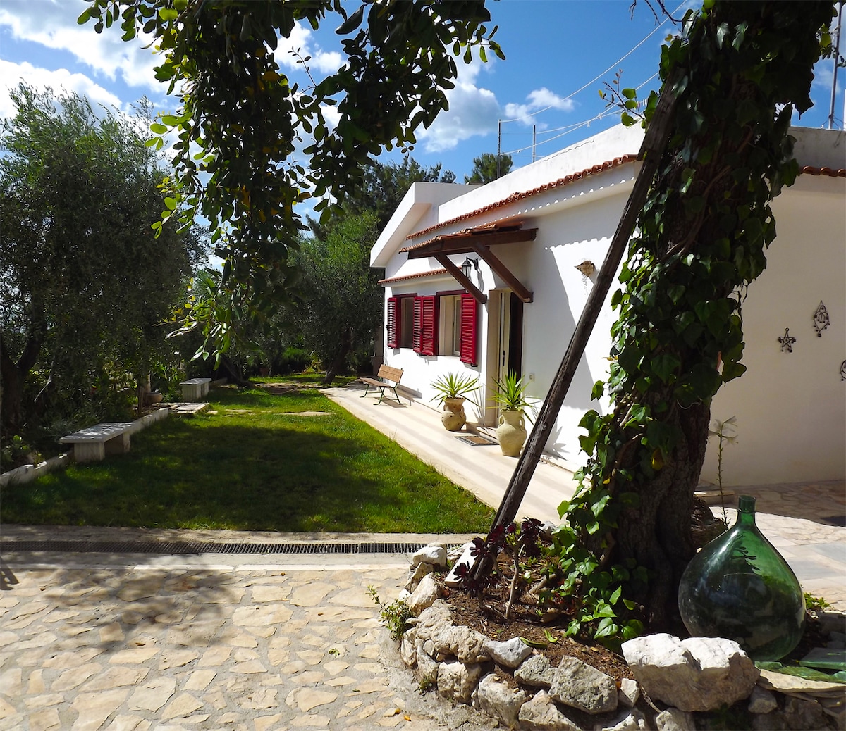 A well-maintained outdoor area is visible, featuring a stone path leading to a white building flanked by green plants and trees. Red shutters frame the windows, offering a cheerful contrast. A spacious lawn is accessible, providing an inviting space for relaxation.