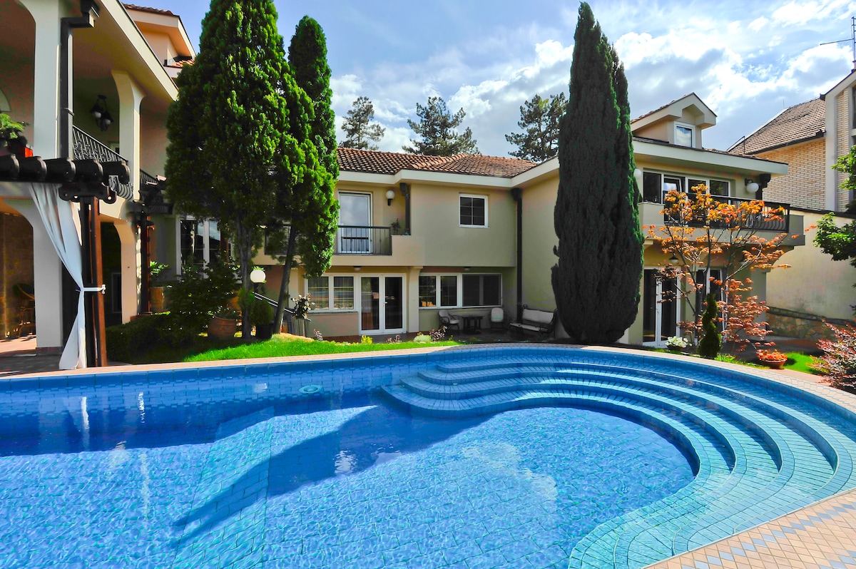 A serene swimming pool is set within a beautifully landscaped area, featuring curved steps leading in. The surrounding greenery includes tall trees and well-maintained shrubs, with a glimpse of the villa's exterior and windows in the background.