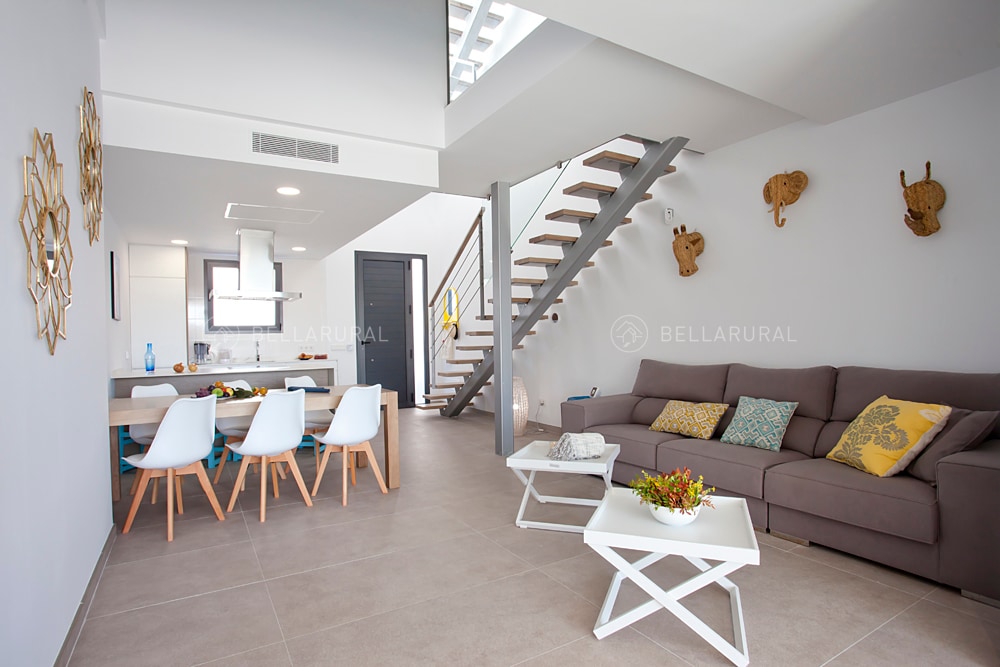 A modern living area features a comfortable sectional sofa with decorative pillows, paired with a minimalist coffee table. A dining space with white chairs surrounds a sleek table, while a stylish staircase leads to the upper floor. Natural light streams through large windows, enhancing the open layout.
