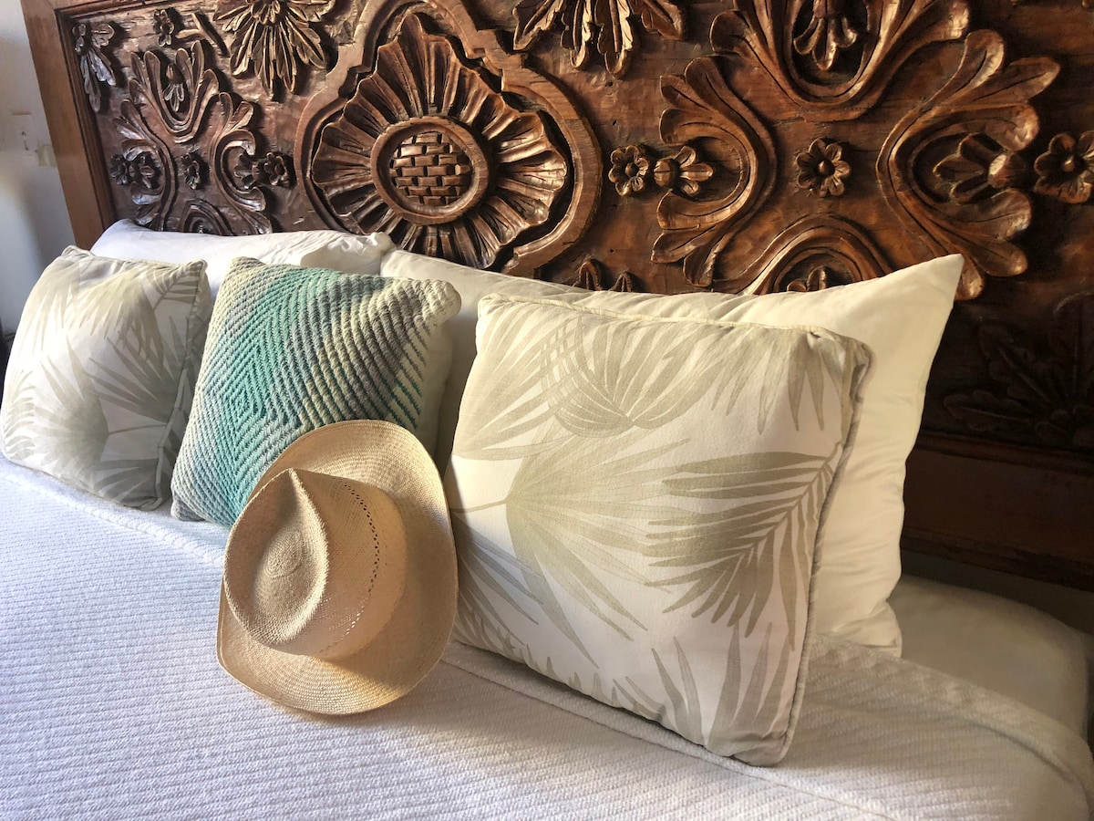 A queen-sized wicker bed is adorned with decorative pillows featuring tropical patterns. A straw hat rests invitingly on the white bedding, complemented by a beautifully carved wooden headboard displaying intricate floral designs.