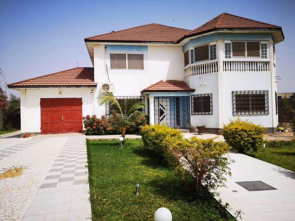 The villa features a two-story exterior with a distinctive design, characterized by a combination of white walls and a terracotta roof. A manicured garden surrounds the entrance, complemented by a stone pathway leading to the front door, and a two-car garage is visible on the left.