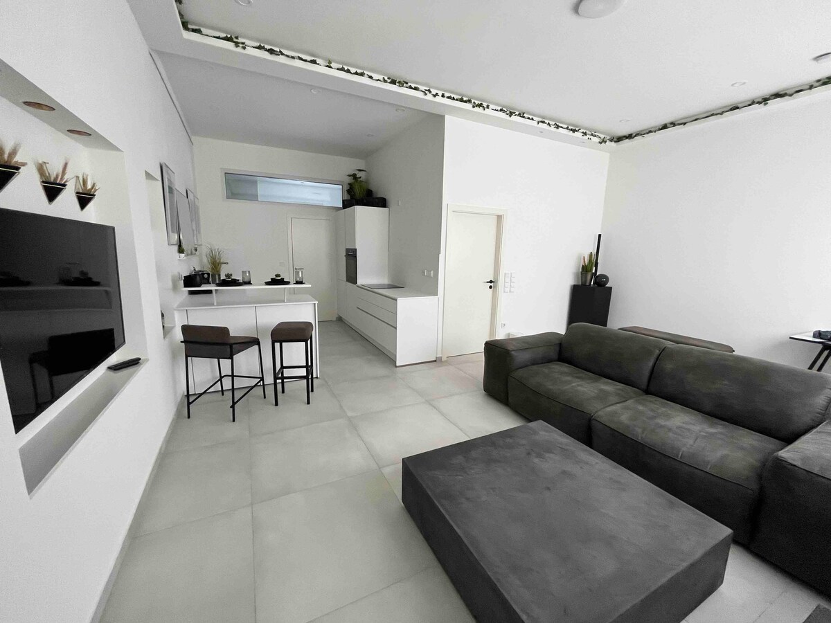 A minimalist living area is showcased, featuring a comfortable dark sofa and a spacious coffee table. A small dining area with bar stools is positioned near the modern kitchen, which contains sleek cabinetry. Natural light filters in through a window, enhancing the open layout.