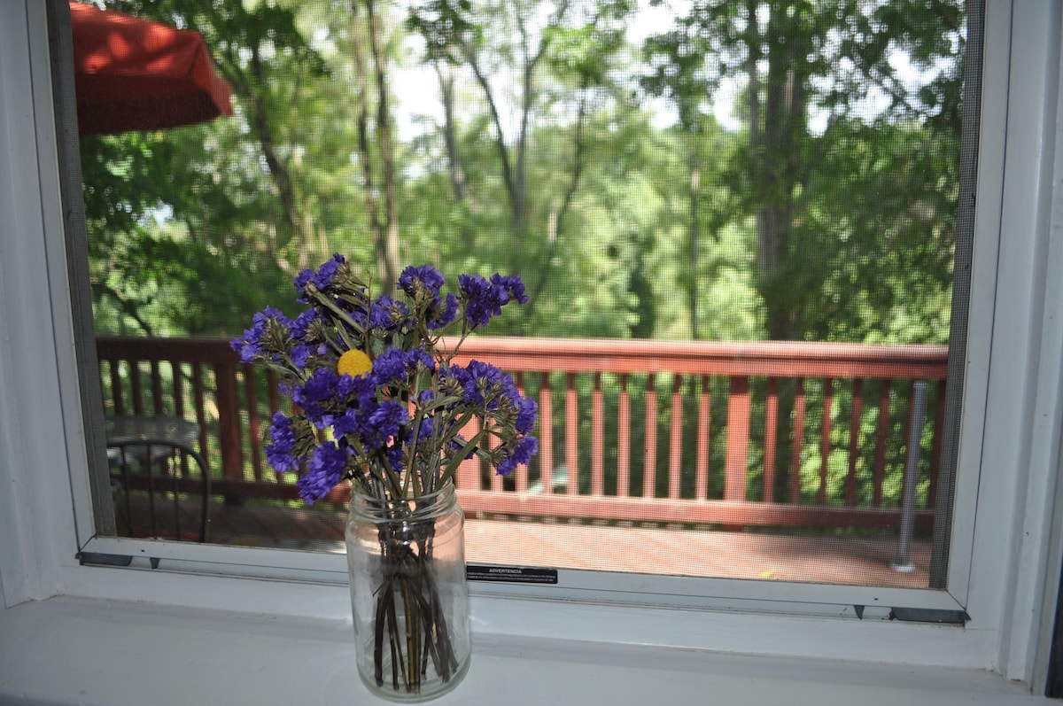 A jar of wildflowers sits on a windowsill, offering a pop of color against the backdrop of a wooded view. A spacious back deck is visible through the window, framed by trees and greenery, suggesting a natural and serene outdoor environment.