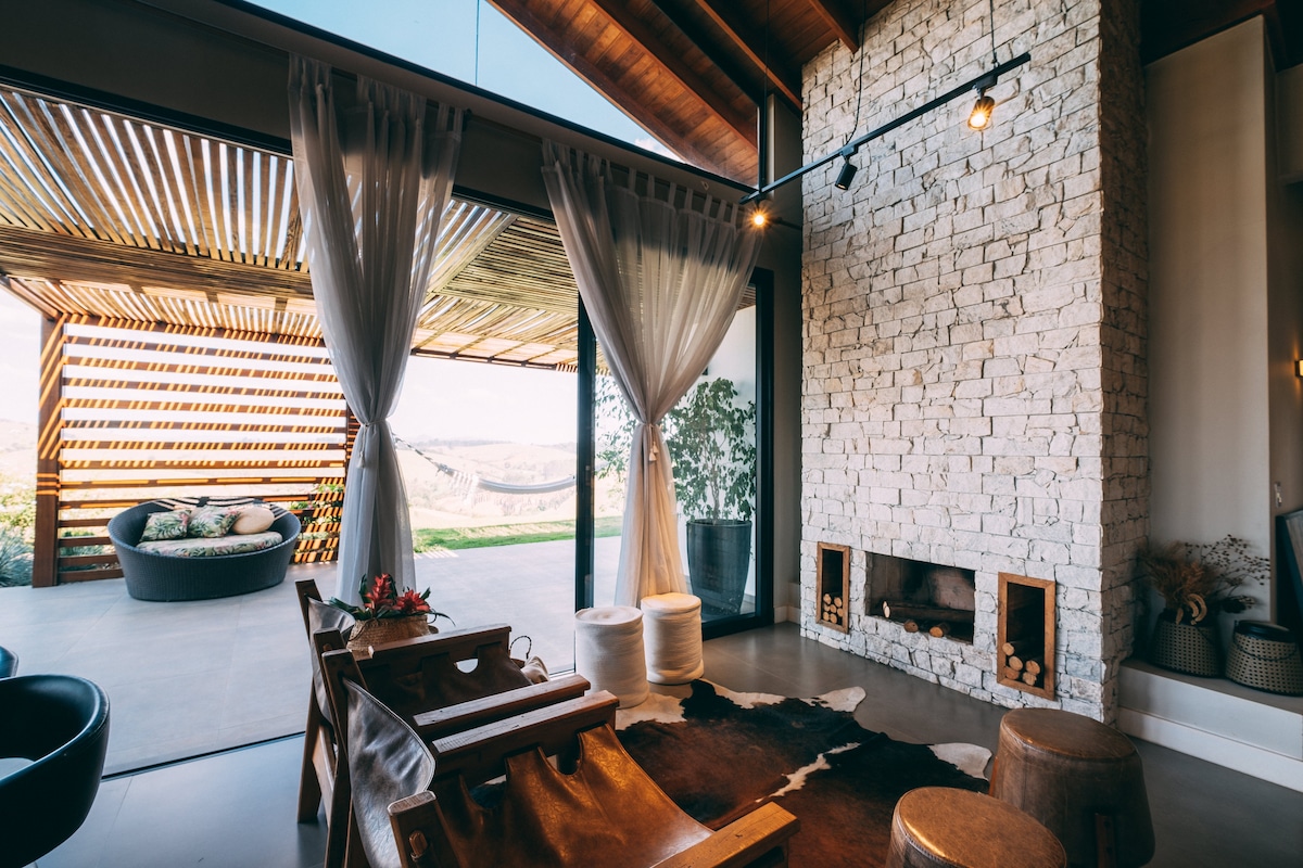 A spacious living area showcases a stone fireplace and large glass doors that open to the outdoors. Natural light fills the room, highlighting a cozy seating arrangement featuring wooden chairs and decorative elements. Soft curtains frame the view of the surrounding landscape.