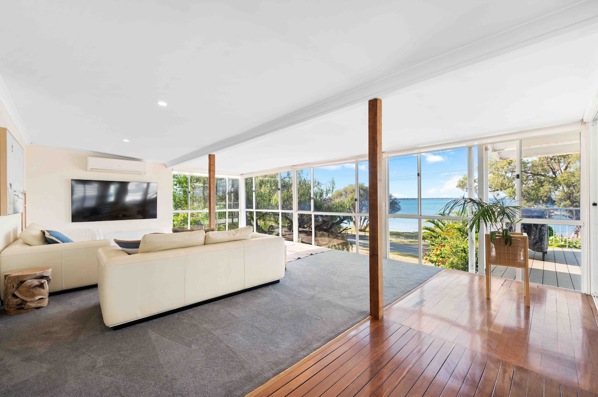 A light-filled living area features large glass doors offering views of the water and surrounding greenery. A spacious white sofa is positioned against one wall, complemented by a flat-screen TV mounted nearby. Natural light enhances the wooden flooring, creating a serene atmosphere.