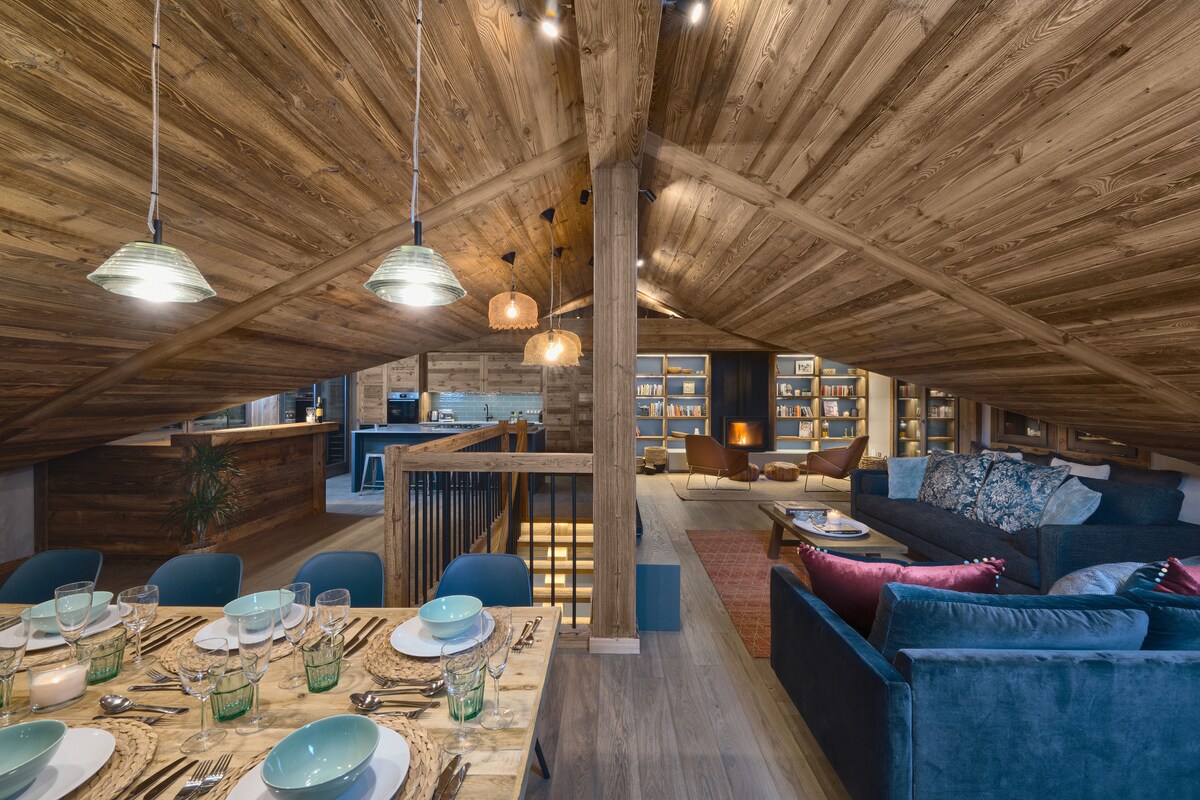 An open-plan upper level features a wooden ceiling with exposed beams. A spacious dining area showcases a large table set with tableware, while a cozy lounge area invites relaxation with plush seating and thoughtfully arranged decor. Warm lighting creates a friendly ambiance throughout.