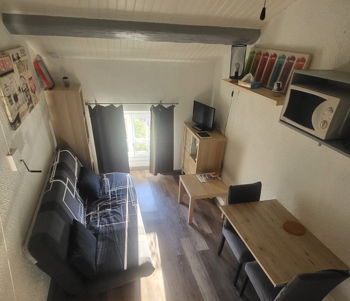 The living area features a modern sofa bed with a geometric pattern, paired with a wooden table and two chairs. A television is positioned on a shelf, while a microwave is installed above. Large windows allow natural light to brighten the space, framed by dark curtains.