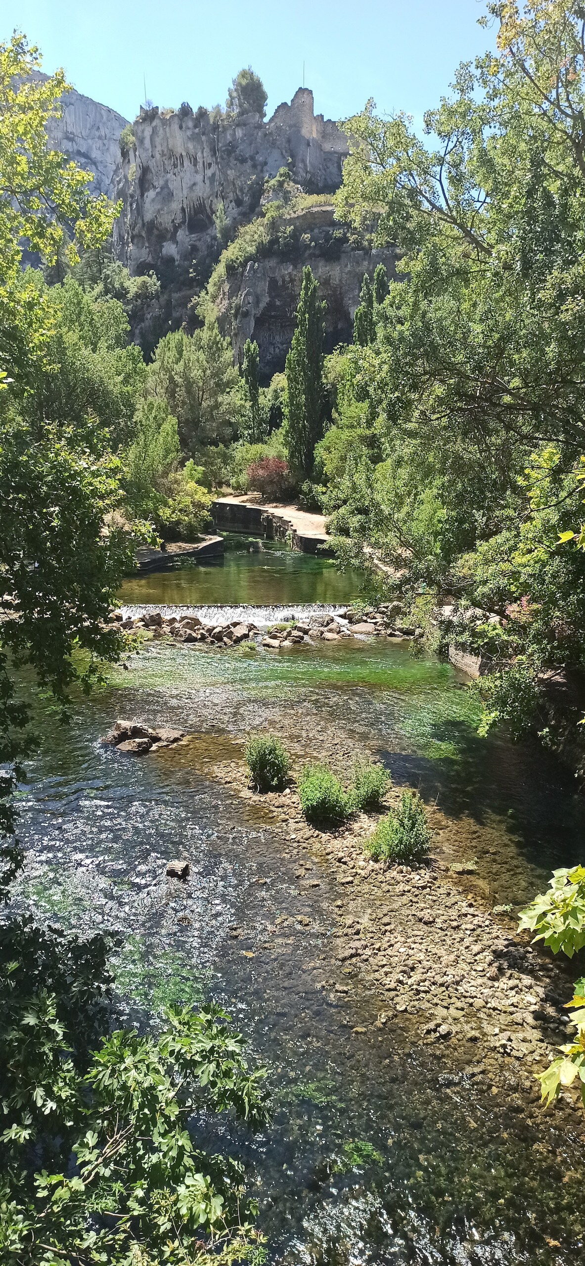 A serene waterway meanders through lush greenery, with gentle ripples visible on the surface. Stone formations and patches of grass are present along the banks. In the background, a rocky hillside is adorned with greenery, adding a natural frame to the tranquil scene.